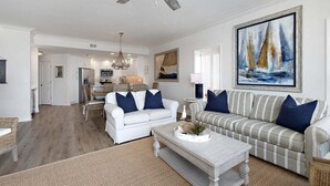 A spacious living room with a white sofa, a striped sofa, and a coffee table. There is a painting of sailboats on the wall. - Luxurious Oceanfront Condominium (St Simons Island)