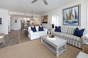 A spacious living room with a white sofa, a striped sofa, and a coffee table. There is a painting of sailboats on the wall. - Luxurious Oceanfront Condominium (St Simons Island)