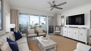 A spacious living room with a large flat-screen TV, a ceiling fan, and a view of the outdoors through a sliding glass door. - Luxurious Oceanfront Condominium (St Simons Island)
