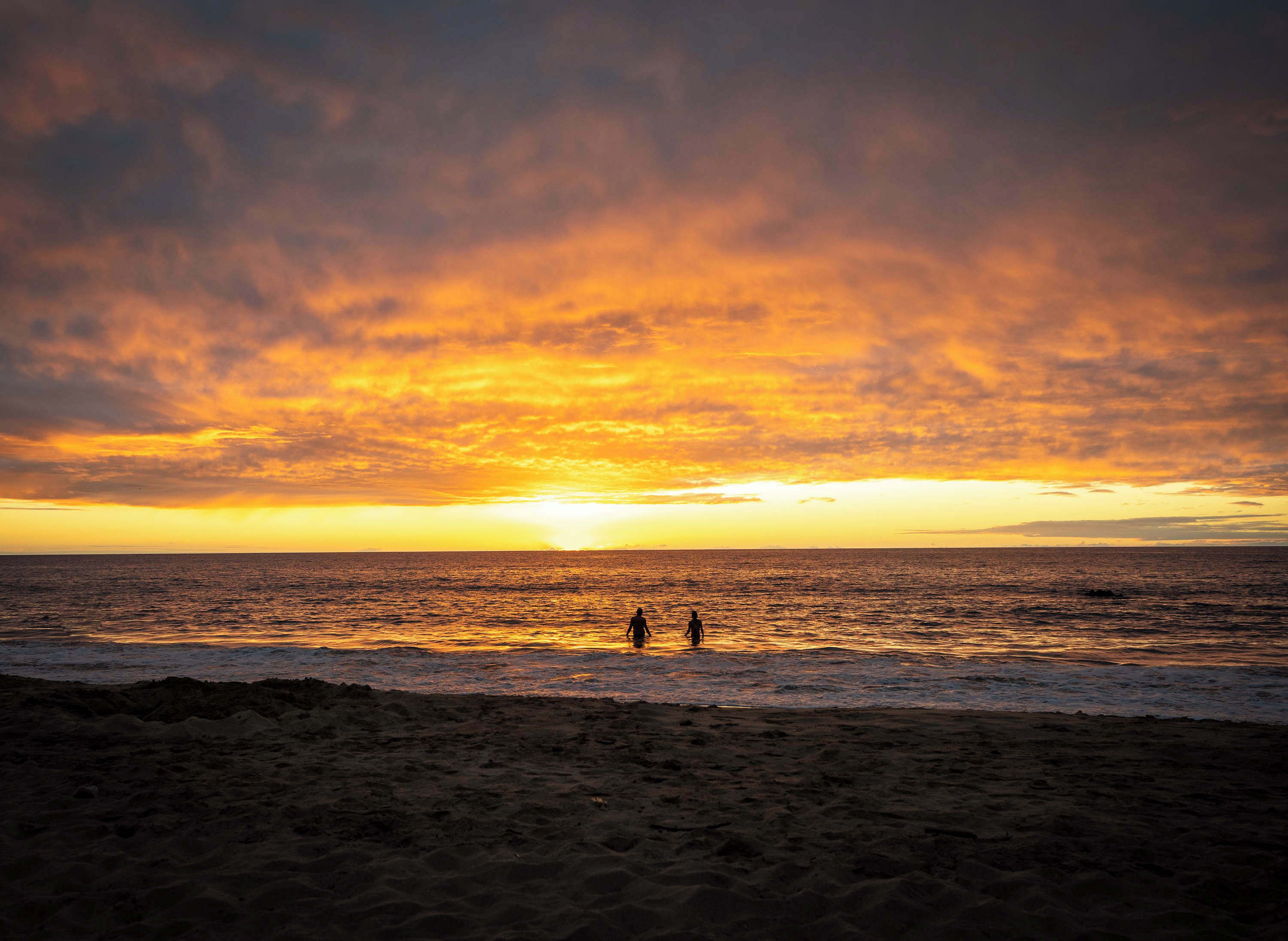 A beach with a sunset over the ocean, two people walking along the shore, and a sandy beach in the foreground.