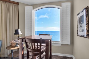 A dining area with a table and chairs, a lamp, a basket of bread, and a view of the ocean through a window with shutters.