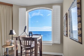 A room with a view of the ocean, a wooden table with chairs, a lamp, and a basket of bread.
