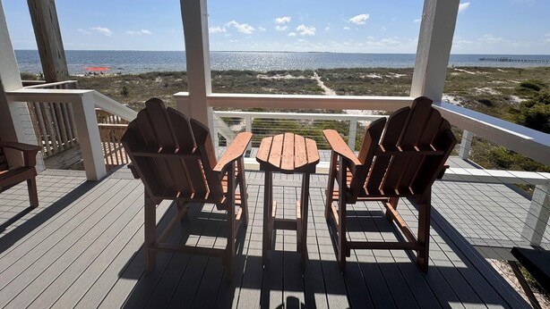 A wooden deck with two Adirondack chairs and a small table, overlooking a beach with a clear view of the ocean and a distant pier. - Fully Remodeled 2025 Beachfront Escape- Pet Friendly- Steps to Sand & Gulf Views (St Joe Beach)