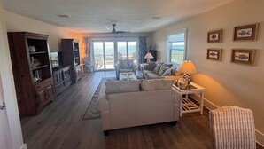 A spacious living room with a sofa, a coffee table, and a dining area with a table and chairs. There is a large window providing a view of the outdoors. - Fully Remodeled 2025 Beachfront Escape- Pet Friendly- Steps to Sand & Gulf Views (St Joe Beach)
