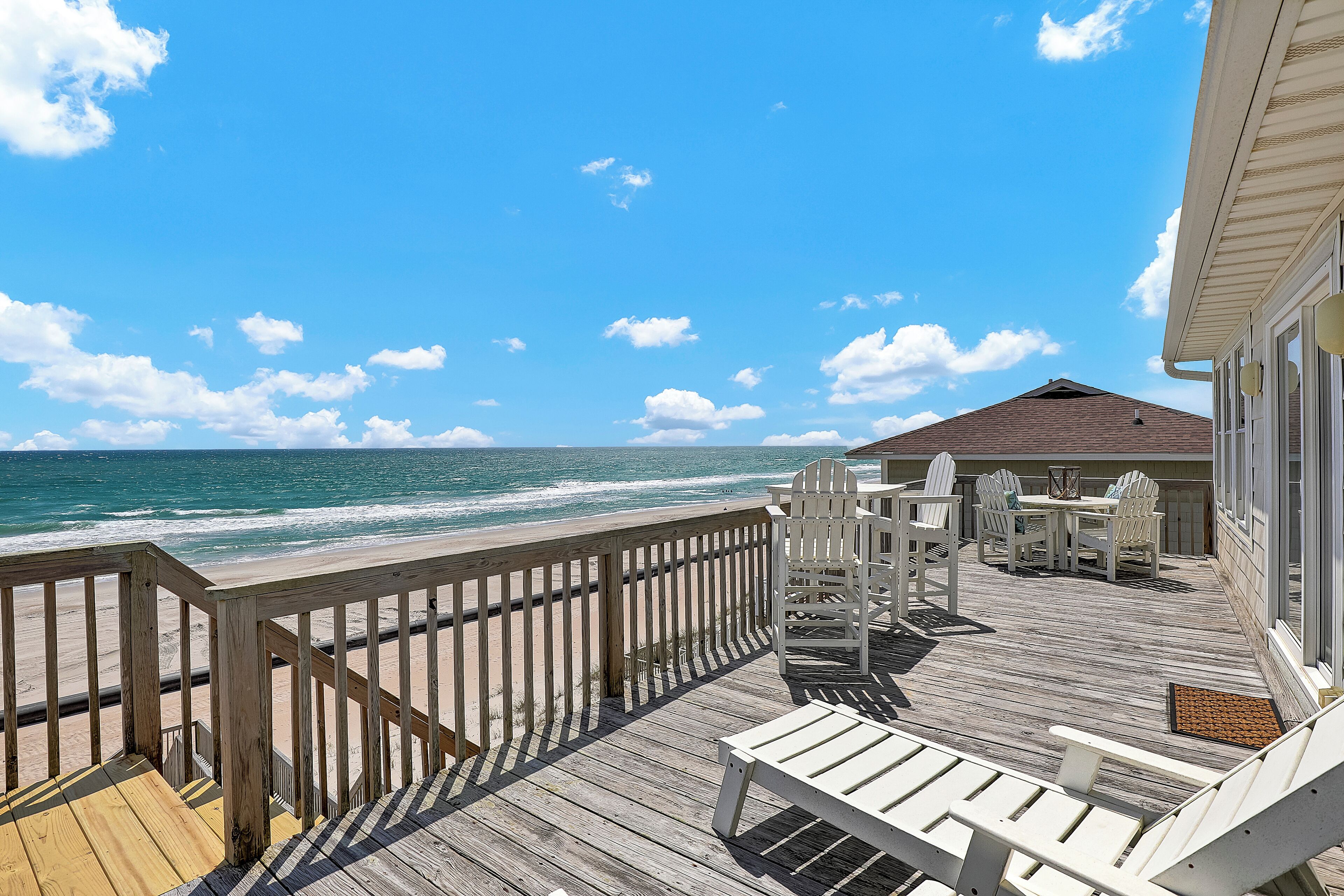 Une terrasse en bois avec des chaises Adirondack blanches et une table, donnant sur une plage où l’on peut admirer les vagues.