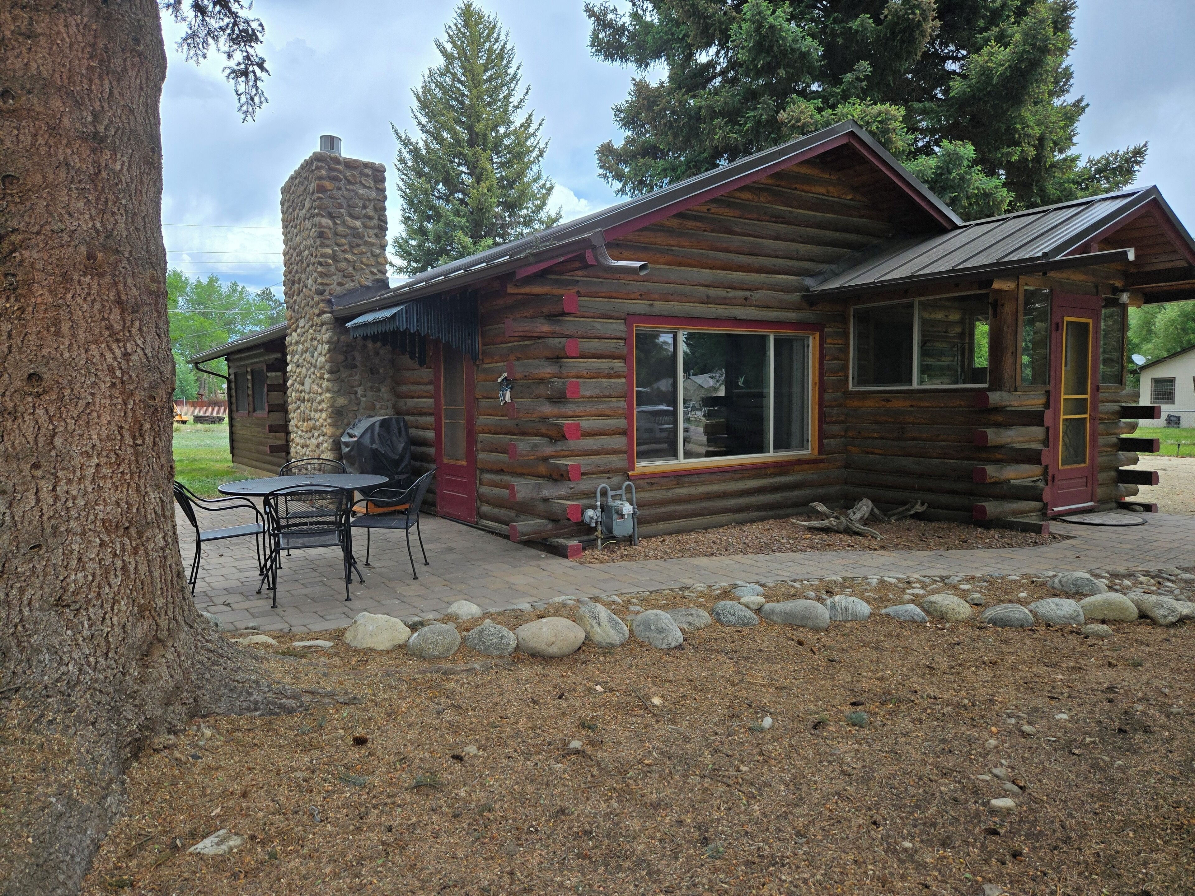 Eine Blockhütte mit einem Steinkamin, einem überdachten Grill und einer Terrasse mit Gartenmöbeln.