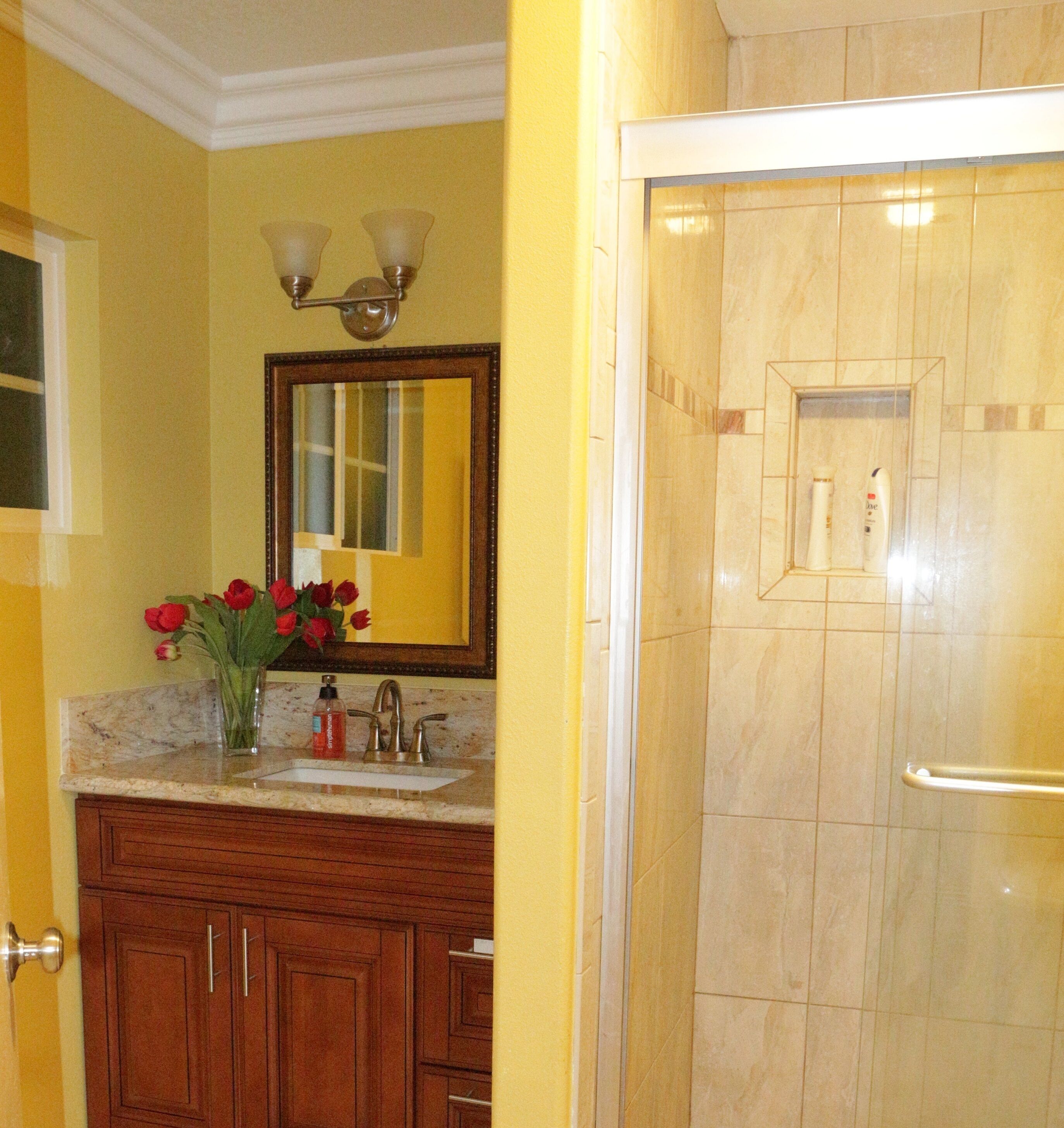 A bathroom with yellow walls, a wooden vanity, a mirror with lights, a sink, and a shower.