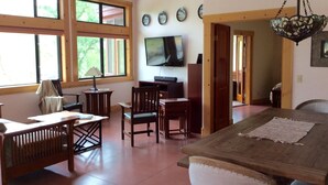 A spacious living room with a wooden dining table, comfortable seating, a television, and large windows allowing natural light.