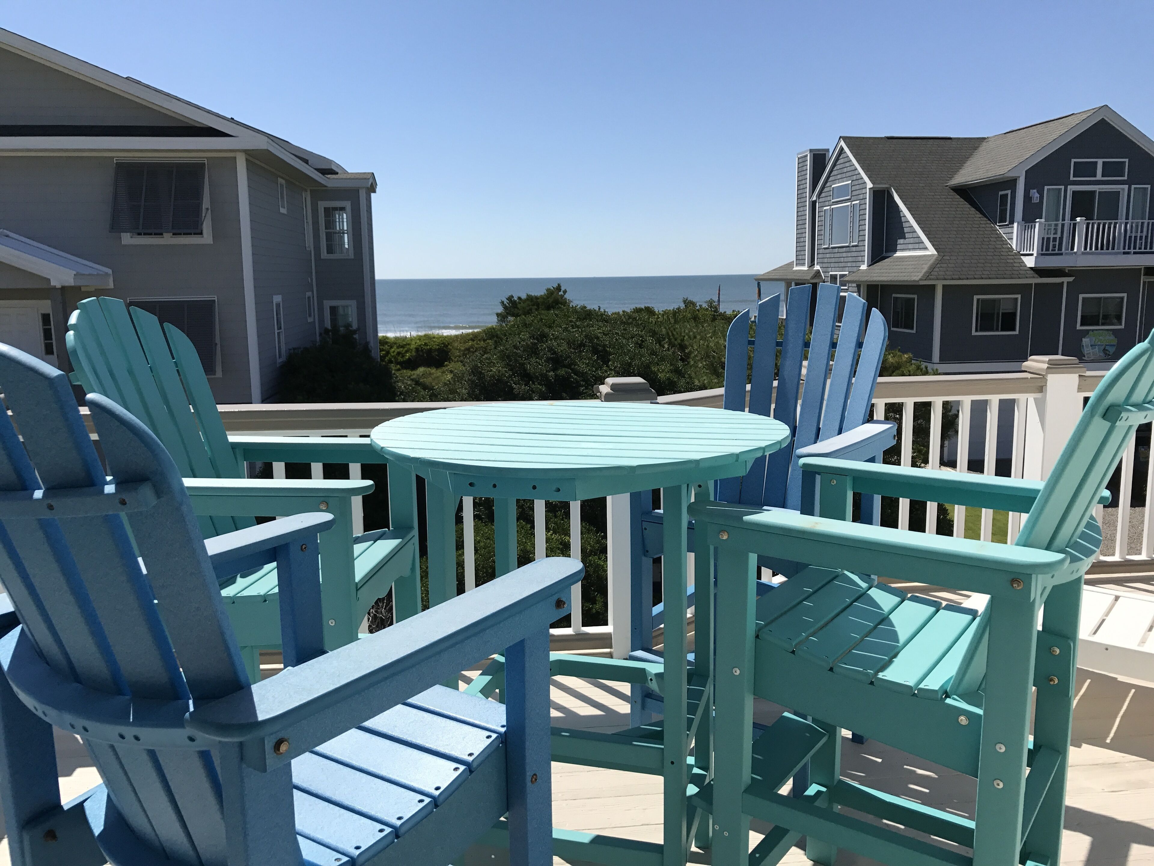 A patio with turquoise Adirondack chairs and a small round table, overlooking a residential area and the ocean.