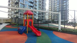 Un terrain de jeux avec un toboggan rouge et une structure d’escalade bleue, entouré d’un immeuble résidentiel à plusieurs étages.