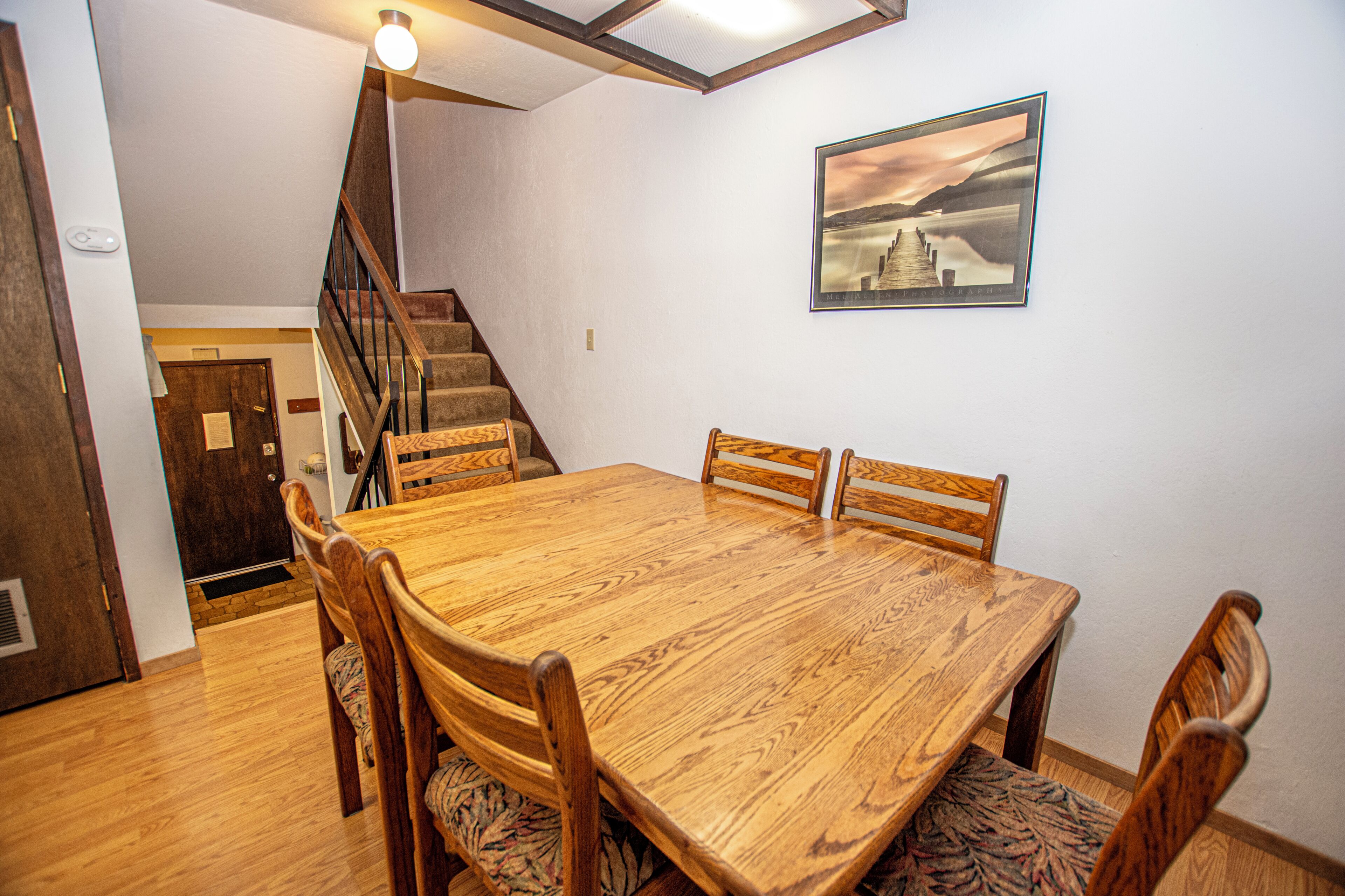 Un comedor con una mesa y sillas de madera, una escalera y un cuadro en la pared.