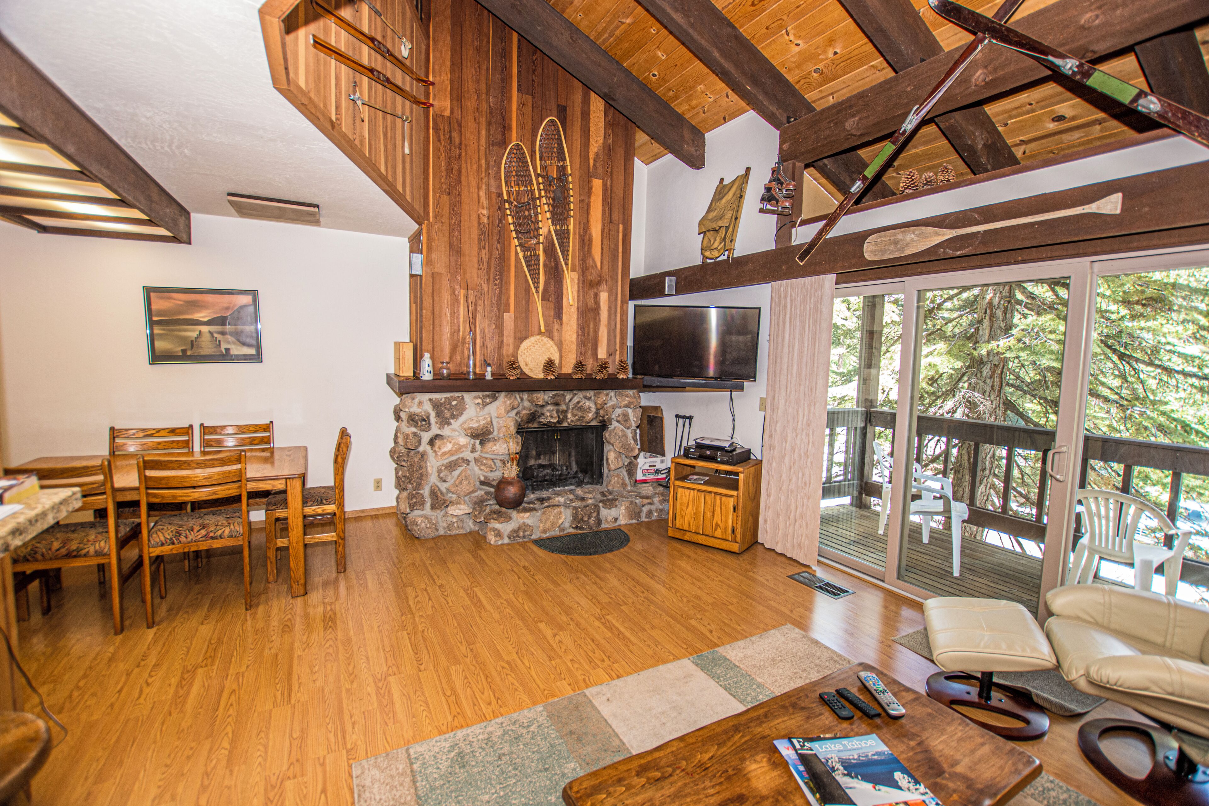 A wooden-floored living room with a stone fireplace, a flat-screen TV, and a large window overlooking a deck.