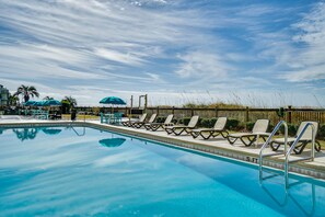 A swimming pool with lounge chairs and umbrellas, surrounded by a wooden fence and palm trees. - Beachfront | 1st Floor Condo | Mariner West | Pool (Panama City Beach)