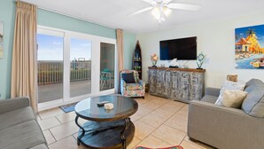 A living room with a large sliding glass door leading to a balcony, a ceiling fan, a flat-screen TV, a round coffee table, and a sofa. - Beachfront | 1st Floor Condo | Mariner West | Pool (Panama City Beach)