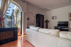 A living room with a white leather sofa, a black entertainment unit, and a TV. There is a window with curtains and a view of the outside.