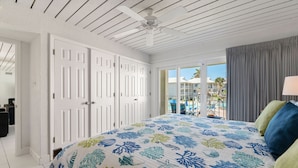 A bedroom with a bed, white doors, a ceiling fan, and a view of a pool and buildings outside.