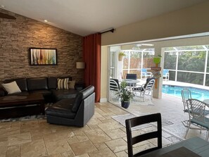 Una sala de estar moderna con una pared de piedra, un sofá de cuero negro, una zona de comedor con sillas blancas y vista a una piscina.