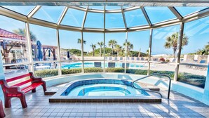 A glass-enclosed indoor swimming pool with a red bench, clear blue tiles, and a view of the beach and palm trees. - Tops'l Resort Beach Manor Ocean Blue Chic Condo! Beachfront with private beach! (Miramar Beach)