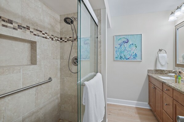 Un baño moderno con ducha de vidrio, una toalla blanca, un lavamanos con encimera de granito y un cuadro en la pared.
