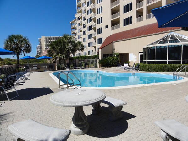 A swimming pool with lounge chairs and umbrellas, surrounded by a building with balconies and a red roof.