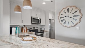 A modern kitchen with white cabinets, stainless steel appliances, a marble countertop, and a large wall clock. - Tops'l Resort Beach Manor Ocean Blue Chic Condo! Beachfront with private beach! (Miramar Beach)