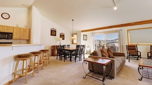 A spacious living area with a sofa, dining table, and bar stools. A kitchen with wooden cabinets and a microwave is visible.