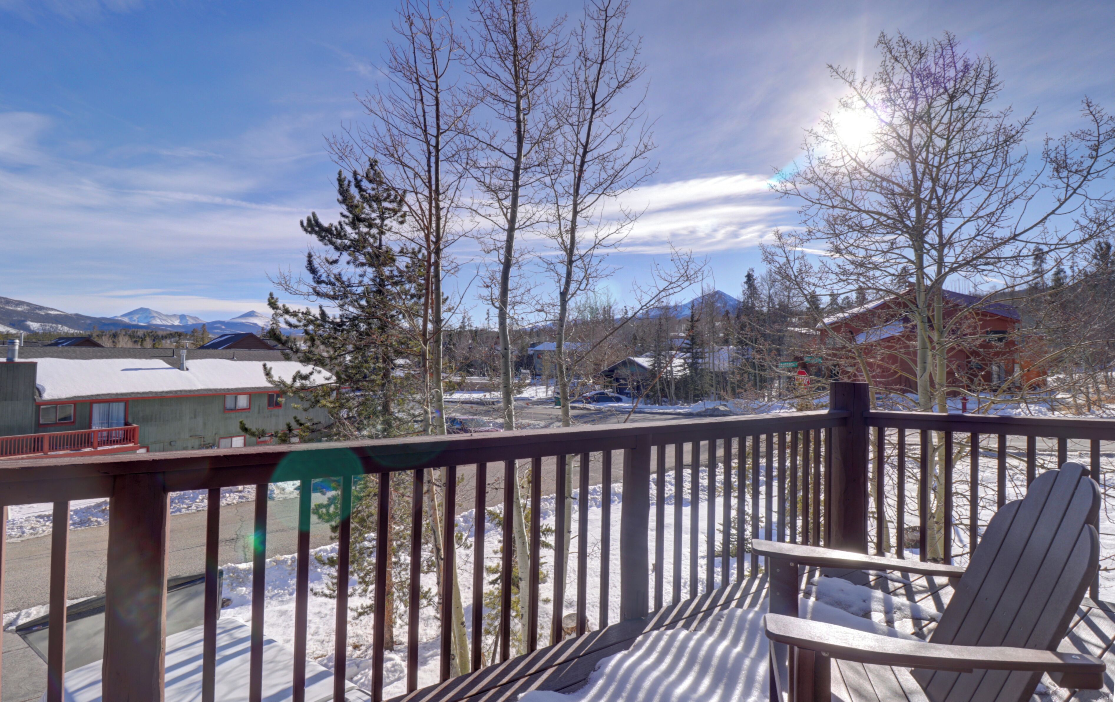 A wooden deck with a chair, overlooking a snowy landscape with buildings and mountains in the background.
