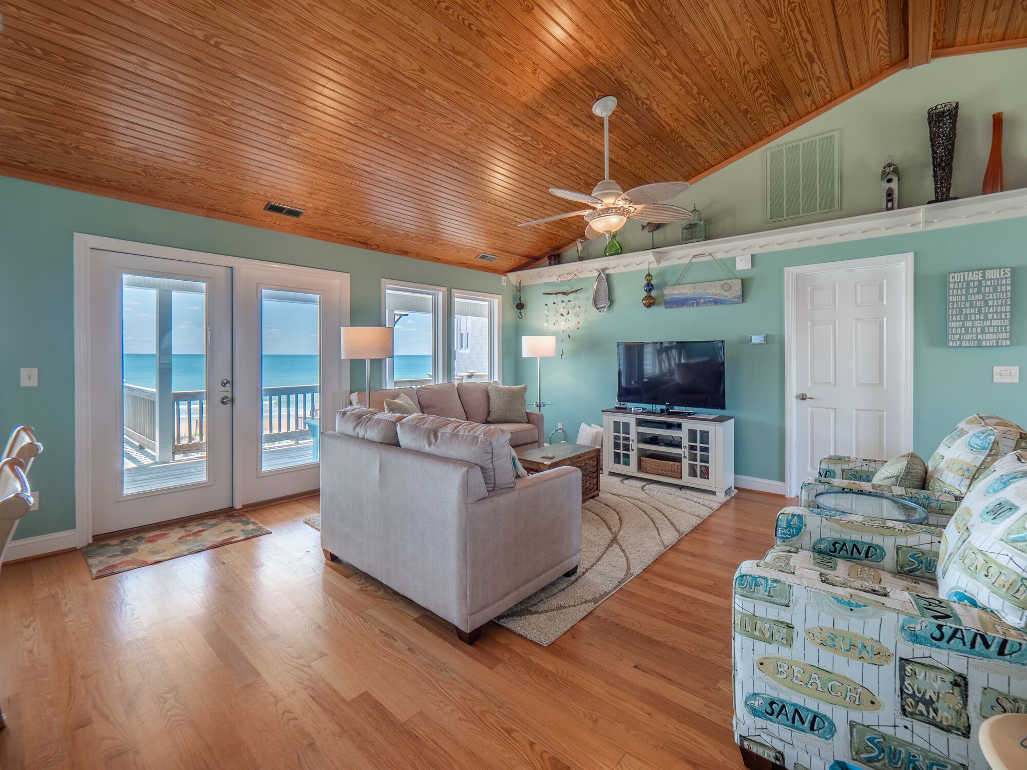 A spacious living room with a large sofa, a television, and a ceiling fan. There are two doors leading to a balcony with a view of the ocean.