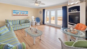 Ein Wohnzimmer mit einem gestreiften Sofa, einem Glastisch, einem Korbstuhl und Blick auf den Strand durch eine Glastür.