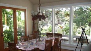 A dining area with a table set for six, surrounded by wooden chairs, large windows offering a view of greenery, and a tripod in the background. - Riverfront Lodge great setting, walk to downtown! (Bend)