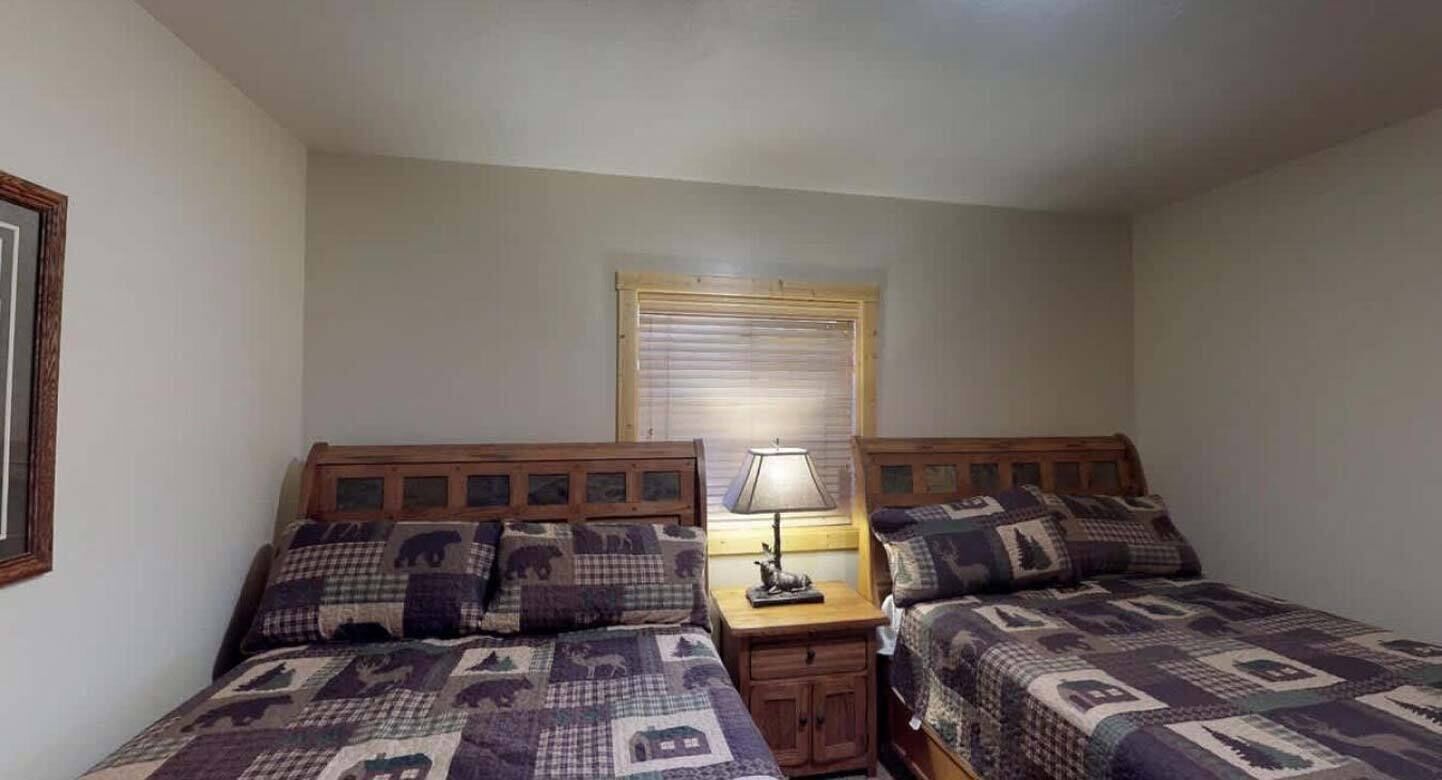 A bedroom with two beds, a nightstand with a lamp, a window with blinds, and a framed picture on the wall.
