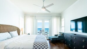 A hotel room with a bed, a ceiling fan, a TV, a dresser, and a view of the ocean through the windows.