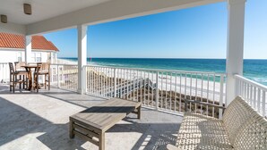 A covered outdoor area with a wooden table and chairs, a white bench, and a view of the beach and ocean.