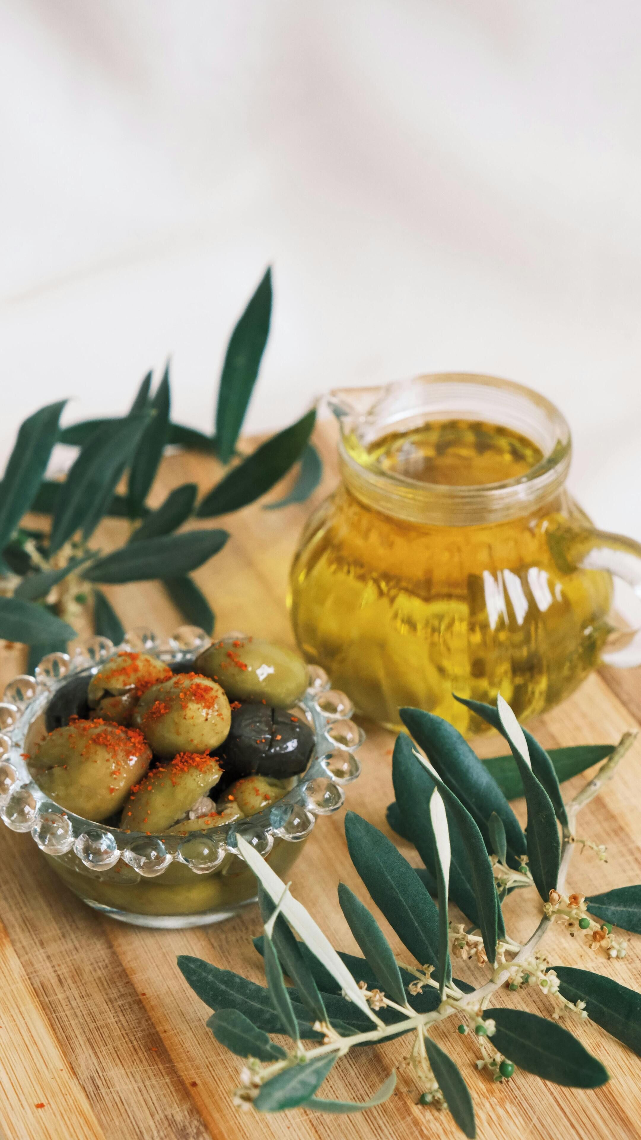 A glass jar of honey, a bowl of olives, and olive branches on a wooden surface.