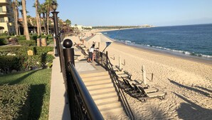 Un paseo marítimo que conduce a una playa arenosa con sillas de descanso y sombrillas, palmeras y una vista despejada del océano.