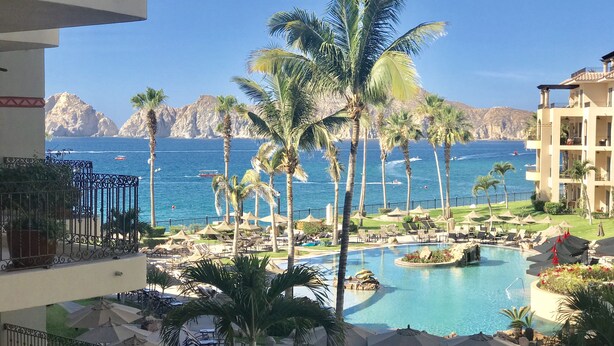 Zona de piscina en un resort con palmeras, sillas de descanso y vistas al mar y las montañas.