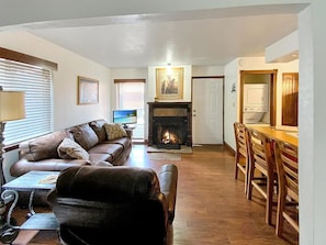 A living room with a fireplace, a sofa, a dining table with chairs, and a kitchen area with appliances. - Location Location Location (Angel Fire)