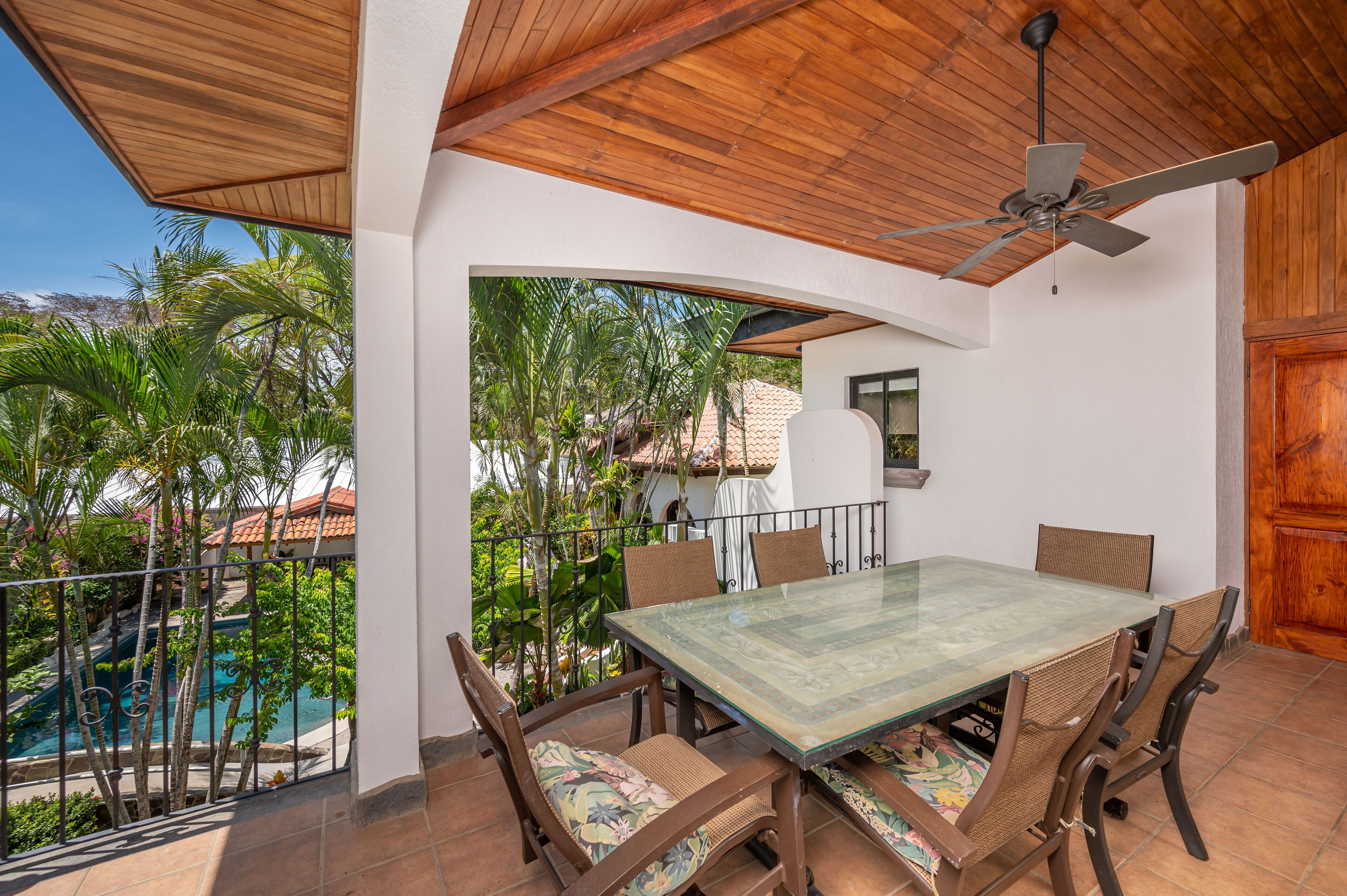 A covered patio with a dining table and chairs, a ceiling fan, and a view of a swimming pool and greenery.