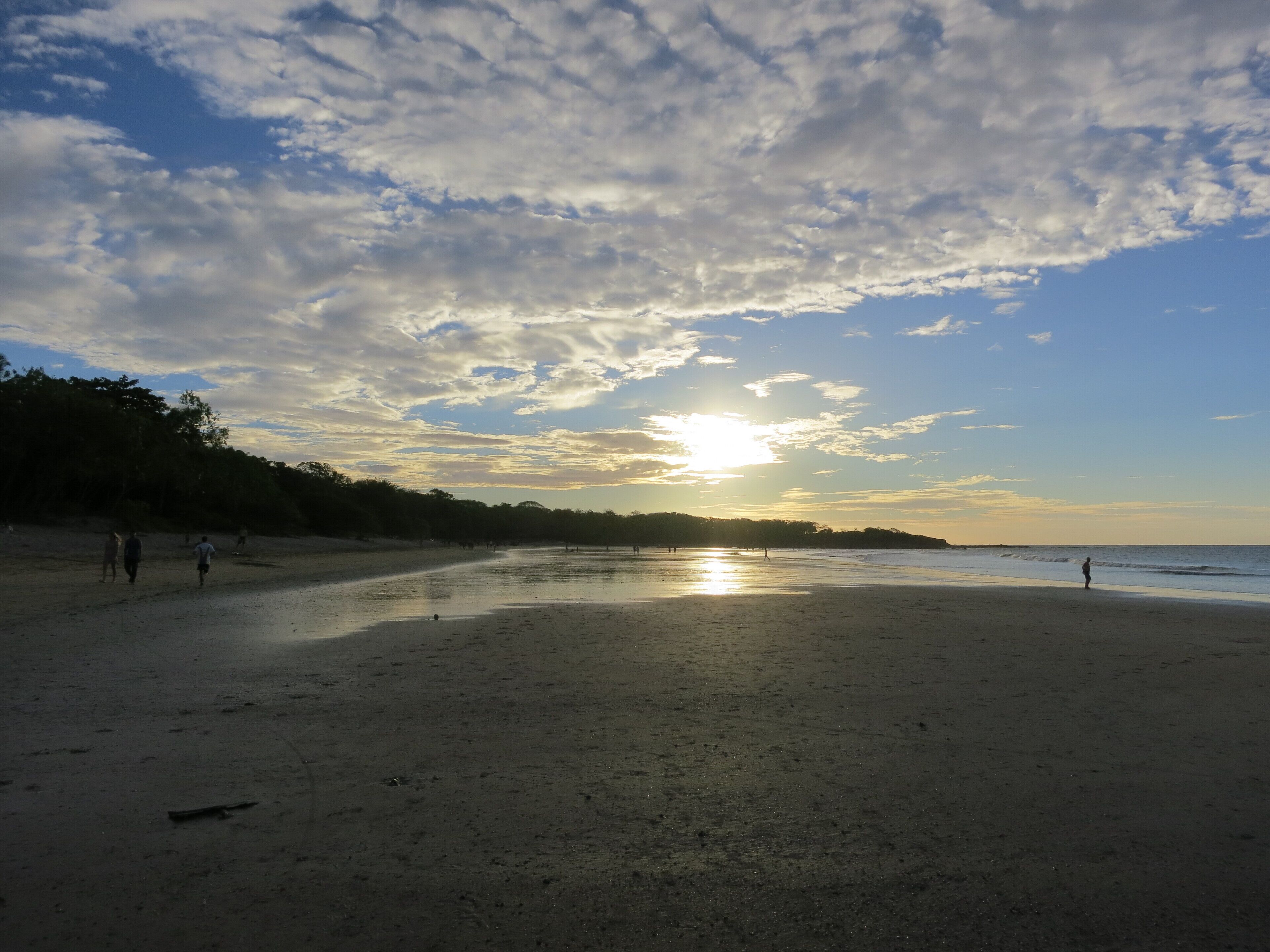 Una playa con un atardecer, gente caminando y árboles al fondo.