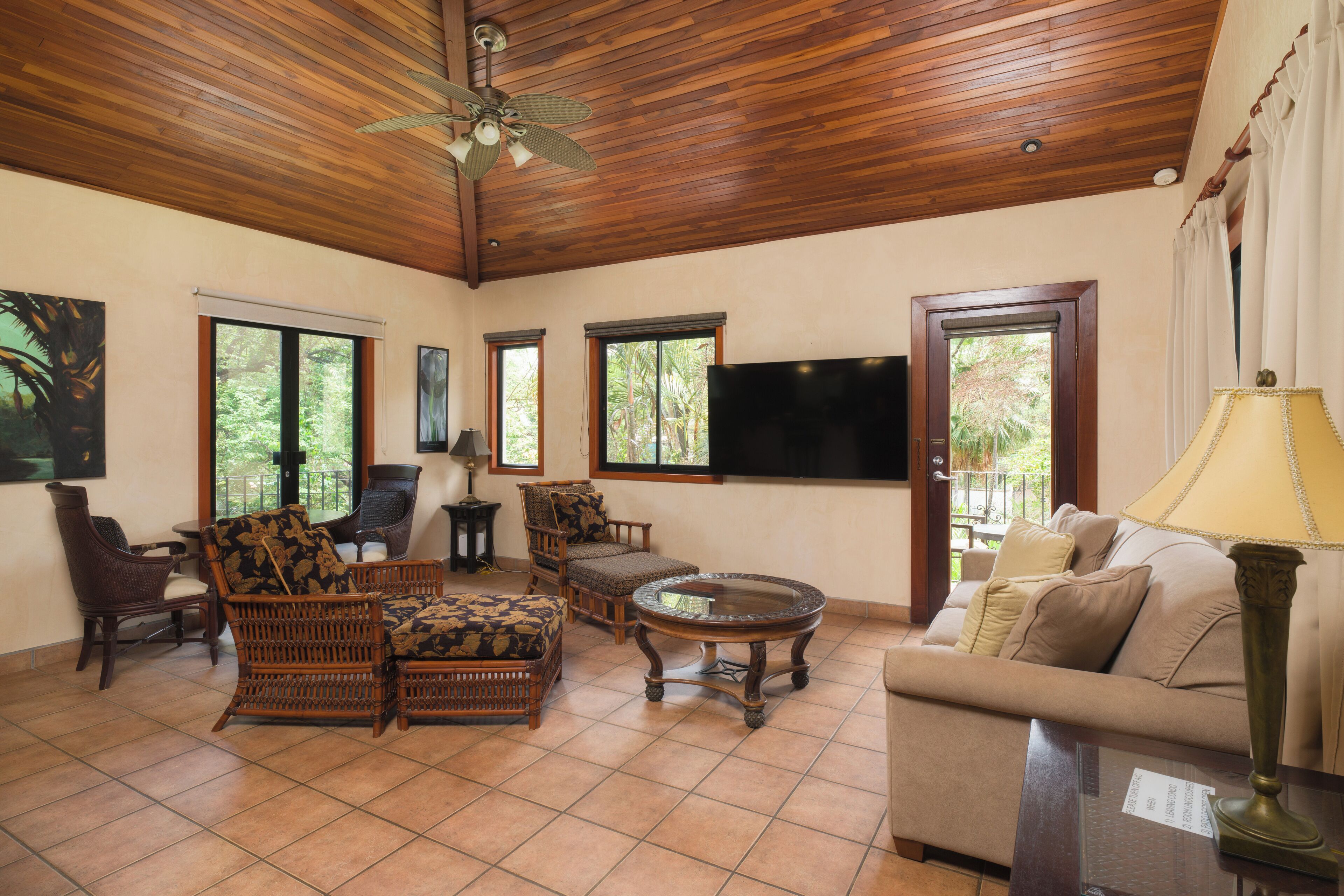 A spacious living area with wooden ceiling, large windows, a flat-screen TV, wicker furniture, and a lamp.