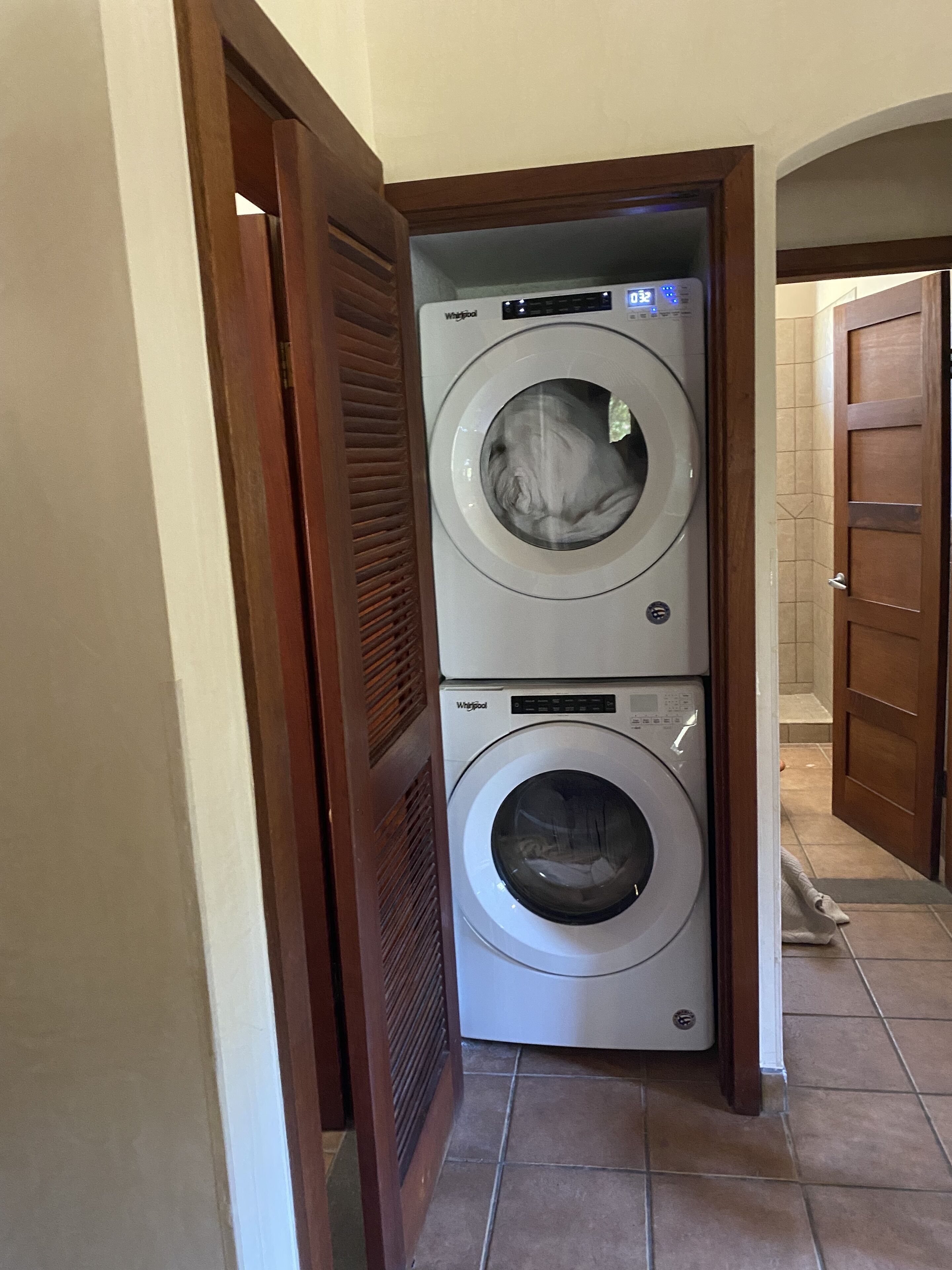 Une machine à laver et un sèche-linge superposés, installés dans un couloir avec un cadre de porte en bois et un revêtement de sol carrelé.