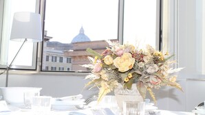 Une table dressée pour le dîner, avec un vase de fleurs au centre, une lampe sur le côté gauche et, par la fenêtre, une vue sur un bâtiment en forme de dôme.
