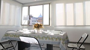 Une salle à manger avec une table dressée pour quatre personnes, un vase de fleurs et une vue sur un bâtiment coiffé d’un dôme, visible à travers une grande fenêtre.