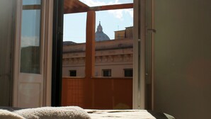 Un lit recouvert d’une couverture blanche et orné d’un coussin à rayures, une fenêtre laissant entrer la lumière du soleil et une vue sur des bâtiments et un dôme en arrière-plan.