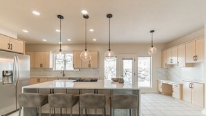 Una cocina moderna con una isla central, taburetes de bar, iluminación colgante y un ventanal grande.
