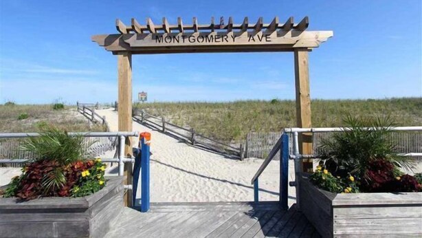 Una pérgola de madera con la inscripción 'MONTGOMERY AVE', que da paso a una zona de playa arenosa con un camino de madera y barandillas.