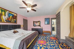 Un dormitorio con cama, ventilador de techo, obras de arte, espejo y una alfombra estampada.