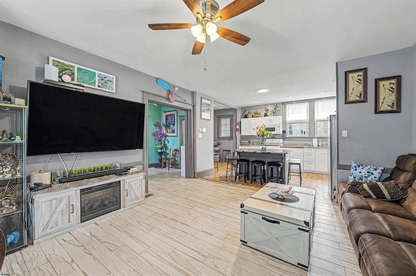 A spacious living area with a large flat-screen TV, a ceiling fan, and a kitchen in the background.