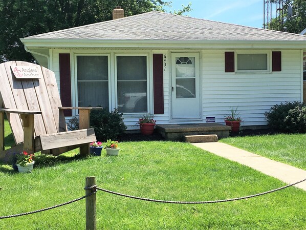 A small, single-story house with a front porch, a wooden chair, and potted plants.
