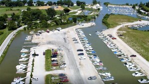 Une vue aérienne d’une marina où des bateaux sont amarrés le long du quai, une route qui longe la marina, et une vue dégagée sur l’océan en arrière-plan.
